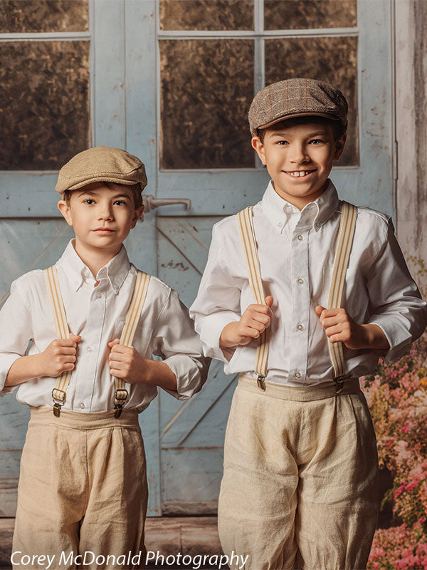 Two young boys wearing white button-down shirts, beige pants, suspenders, and flat caps stand side by side holding their suspenders in front of a rustic blue wooden door backdrop with window panes and soft floral accents.