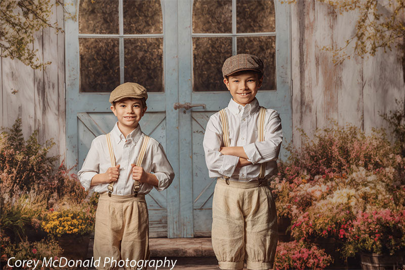 Two young boys wearing white button-down shirts, beige shorts with suspenders, and flat caps stand in front of a rustic blue wooden double door backdrop surrounded by potted flowers and soft garden foliage, one smiling while the other stands with arms crossed.