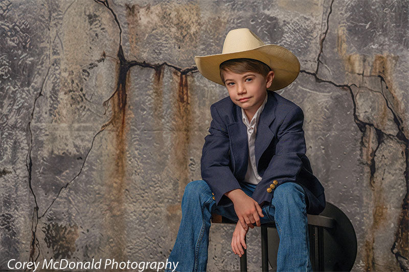 Young boy wearing a navy blazer, white shirt, jeans, and a cowboy hat sits on a bench in front of a rustic cracked concrete wall backdrop with gray industrial texture.