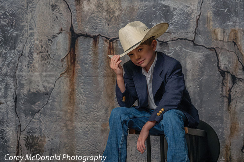 Young boy wearing a navy blazer, white shirt, blue jeans, and a tan cowboy hat sits on a bench leaning slightly forward while touching the brim of his hat in front of a rustic cracked concrete wall backdrop with industrial texture.
