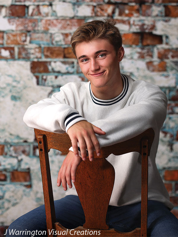 Teen boy leaning on vintage wooden chair in front of a rustic exposed brick and white plaster backdrop, ideal for senior portrait photography.