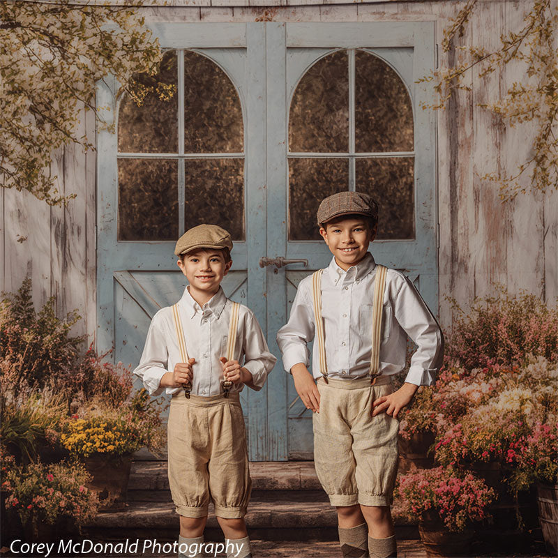 Two young boys in white shirts, beige shorts, suspenders, and caps stand in front of a rustic blue weathered wood door backdrop with visible window panes and colorful flower pots arranged along the base.