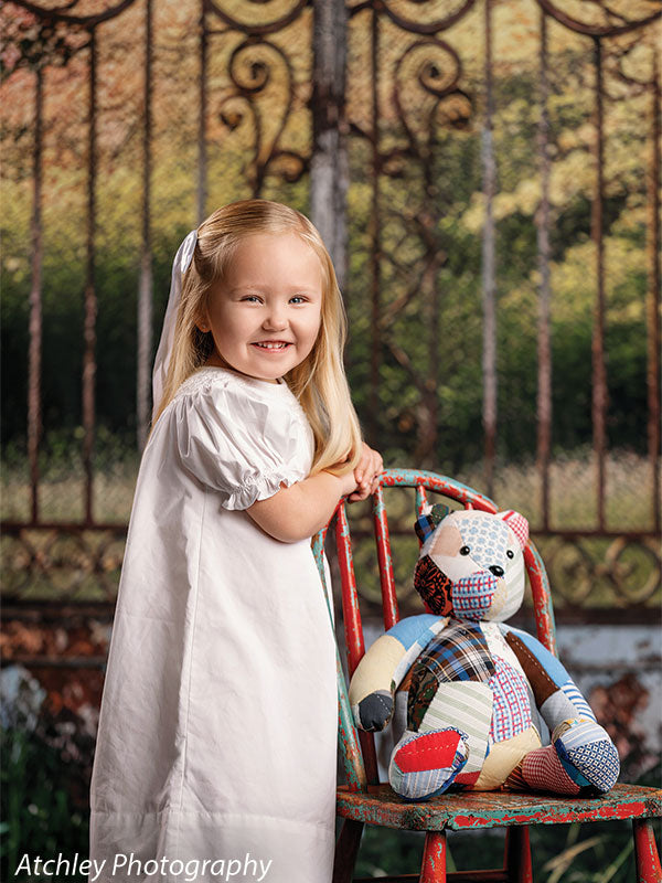 A smiling young girl in a white dress stands next to a small chair with a stuffed teddy bear, set against a rustic garden gate backdrop with weathered iron details.
