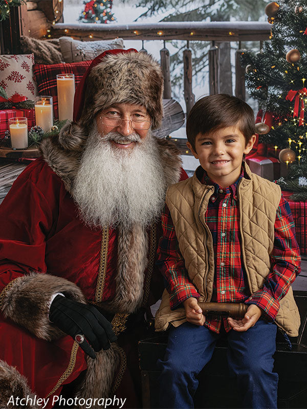 Close view of Santa Claus in a fur hat and red coat smiling next to a young boy in a red plaid shirt and tan vest, seated on a wooden porch with lit candles and a decorated Christmas tree beside them, set against a rustic log cabin holiday backdrop with snowy forest scenery.
