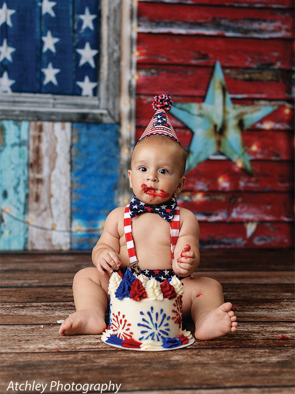 Baby wearing a red white and blue striped party hat with star bow tie and suspenders sitting on a wooden floor in front of a rustic wooden American flag wall backdrop with a large teal star, eating a red white and blue cake decorated with firework patterns.