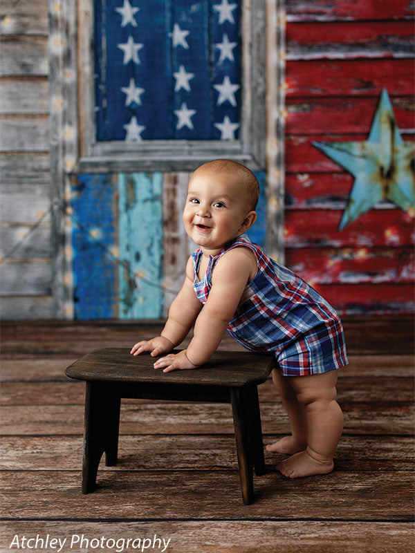 Smiling baby in a red and blue plaid romper standing barefoot while holding onto a small dark wooden stool, posed in front of a rustic wooden American flag wall backdrop with weathered red white and blue planks and a large teal star.