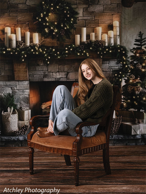 Rustic stone fireplace Christmas backdrop with garland, holiday candles, and festive wreath, shown with a young woman seated in a vintage chair, perfect for cozy holiday portrait sessions.