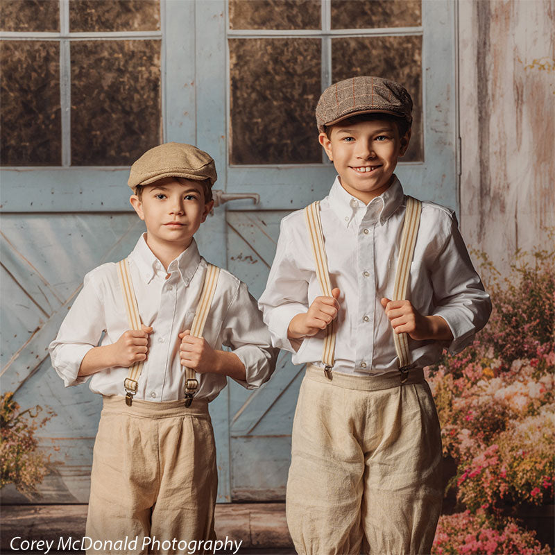 Two young boys wearing white shirts, beige pants, suspenders, and caps stand together smiling and holding their suspenders in front of a rustic vintage blue wooden door backdrop with window panels and surrounding floral details.