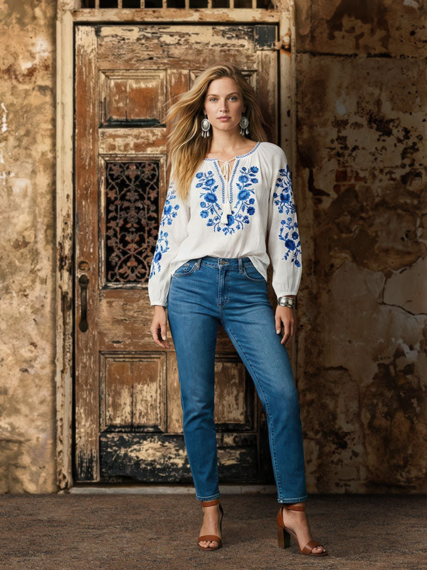 Woman wearing a white blouse with blue embroidered floral details and blue jeans stands with one leg slightly forward in front of a rustic weathered wooden door backdrop with peeling paint and textured stone walls.