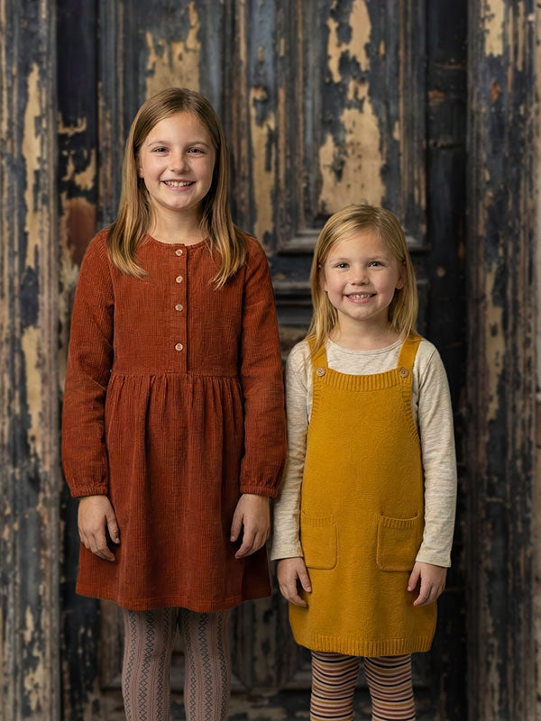 Two young girls stand side by side, one wearing a rust-colored dress and the other wearing a mustard yellow pinafore, posed in front of a dark rustic wooden door backdrop with distressed paint and aged textures.