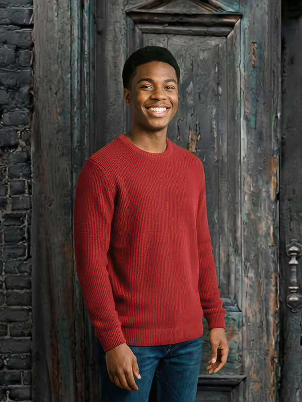 Young man wearing a red sweater and jeans stands smiling with relaxed posture in front of a rustic weathered wooden door backdrop with peeling paint and exposed brick.