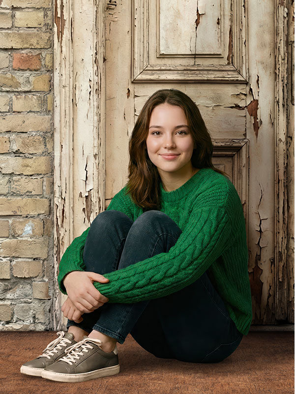 Teenage girl wearing a green sweater and dark jeans sits with knees drawn up, smiling softly in front of a rustic weathered wooden door backdrop with chipped paint and textured brick.