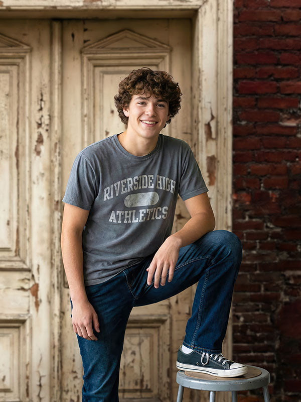 Teenage boy wearing a gray graphic t-shirt and jeans poses with one foot on a stool, smiling in front of a rustic weathered wooden door backdrop with distressed paint and exposed brick details.