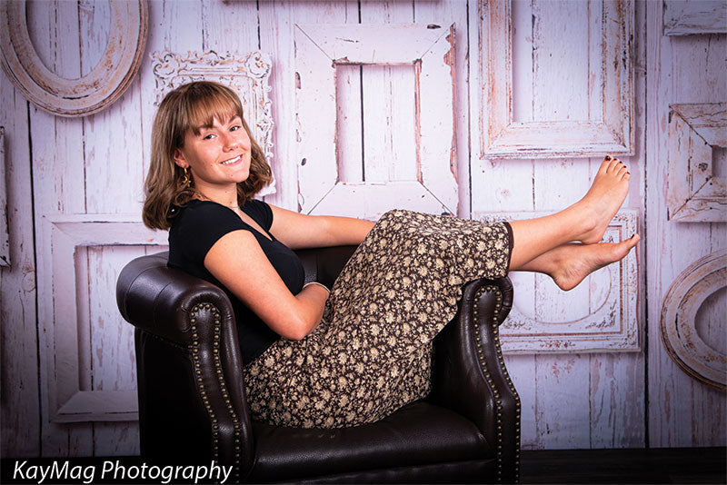 Teen girl reclining on a leather chair in front of a vintage-style backdrop with overlapping whitewashed wood frames, great for editorial or senior session photography.