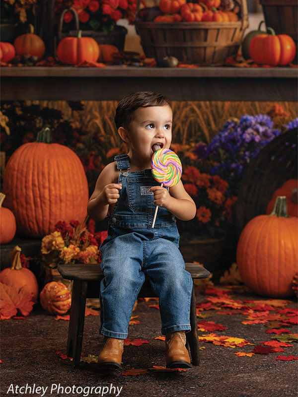 Toddler wearing blue denim overalls and brown boots sitting on a small rustic wooden kids bench while licking a colorful swirl lollipop, surrounded by pumpkins and scattered autumn leaves on a textured dirt floor, with a nostalgic fairground carousel backdrop featuring baskets of gourds and fall flowers behind him.