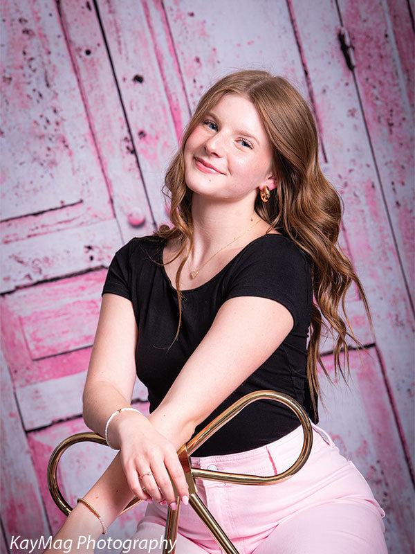 Teen girl seated on a gold chair posing in front of a distressed pink door backdrop, perfect for modern senior portraits with rustic charm.
