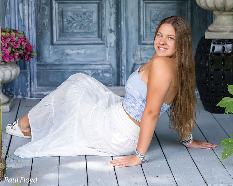 Slate Chateau photography backdrop showing distressed blue-grey carved double doors with pink blossoms, shown with a senior girl in a white outfit seated on the plank floor reclining on one hand CP-8792