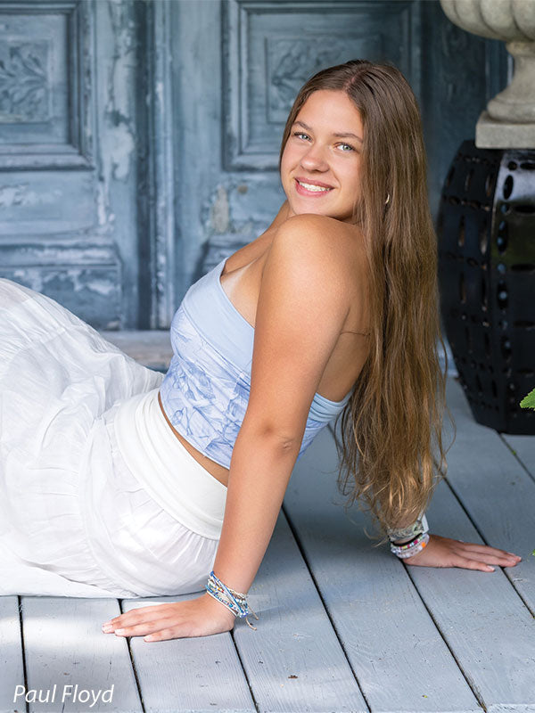 Slate Chateau photography backdrop showing distressed blue-grey carved double doors with pink blossoms, shown with a senior girl in a white outfit seated on the plank floor leaning back on one hand CP-8792