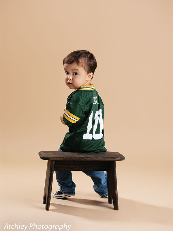 Toddler in a green football jersey with the number 10 sitting sideways on a small rustic wooden bench, turning to look back toward the camera, set against a smooth khaki brown studio backdrop.