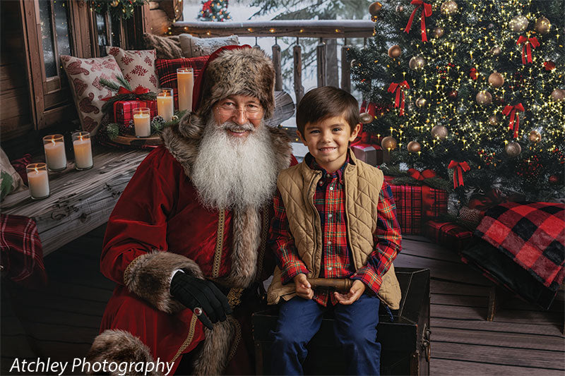 Santa Claus and a smiling young boy sitting together on a wooden porch with red plaid blankets and glowing candles, next to a Christmas tree decorated with gold ornaments and red ribbons, in front of a snowy log cabin Christmas backdrop with pine trees.