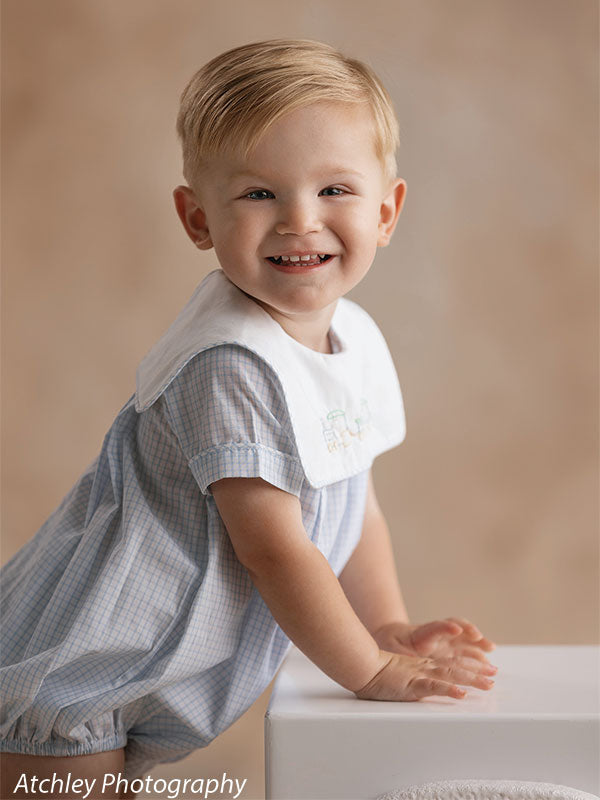 A smiling toddler wearing a light blue romper leans forward with hands on a white cube, posed against a softly textured beige studio backdrop.
