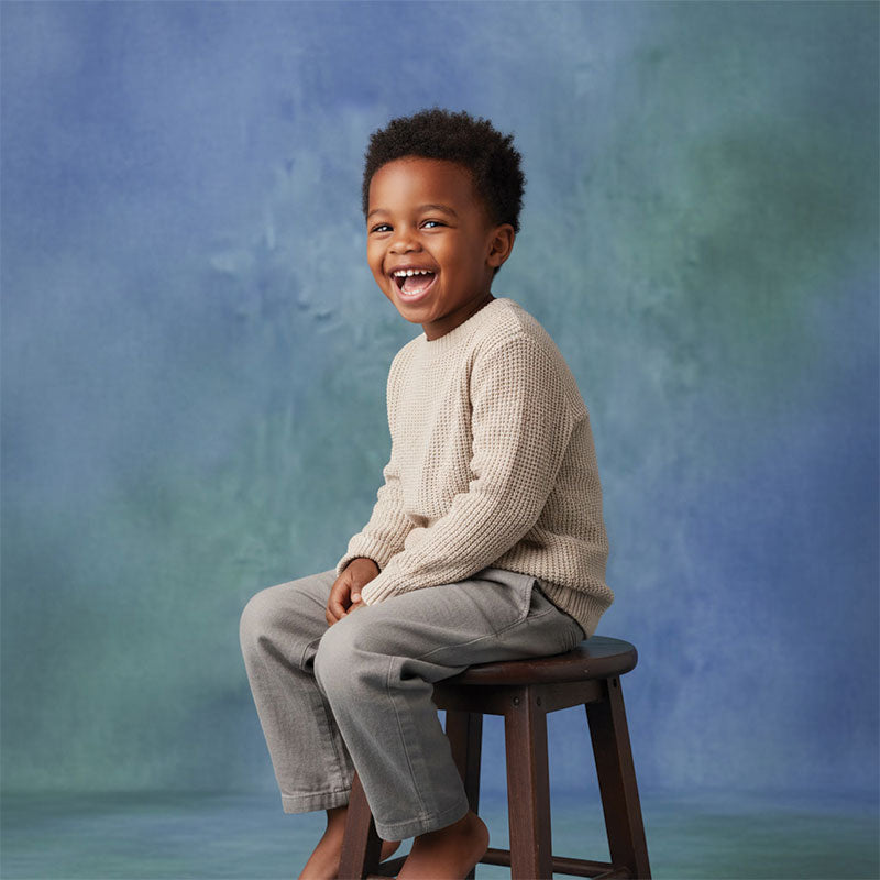 Happy young boy with curly hair wearing a beige knit sweater and gray pants sits on a stool laughing in front of a soft blue and green textured photo backdrop.