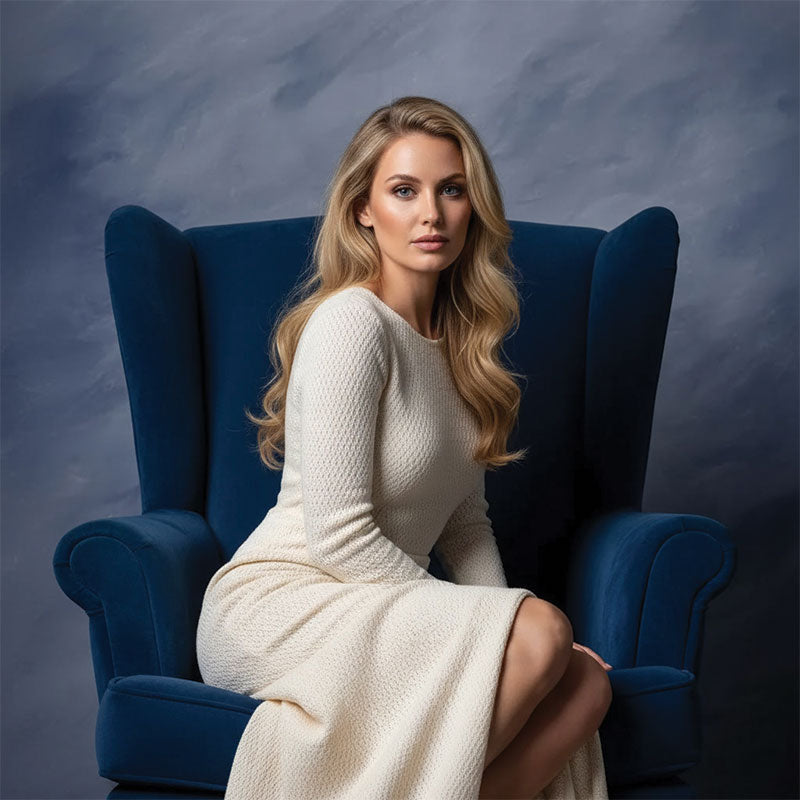 Blonde woman in a cream knit dress sits upright on a blue upholstered armchair looking forward against a soft gray painted portrait backdrop.