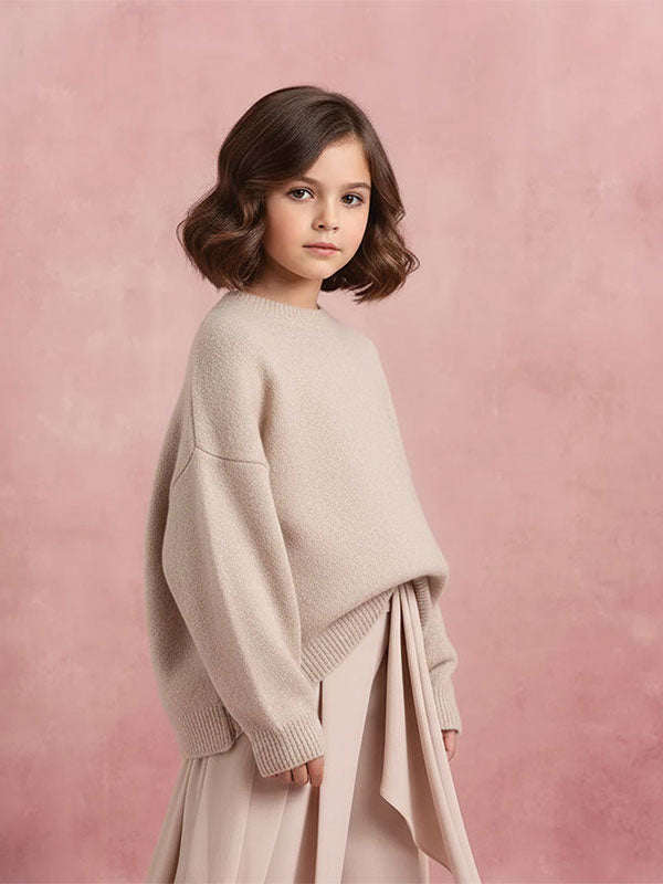 Young girl with short wavy brown hair wearing a beige oversized sweater and flowing skirt stands slightly turned in front of a soft pink mottled studio backdrop.