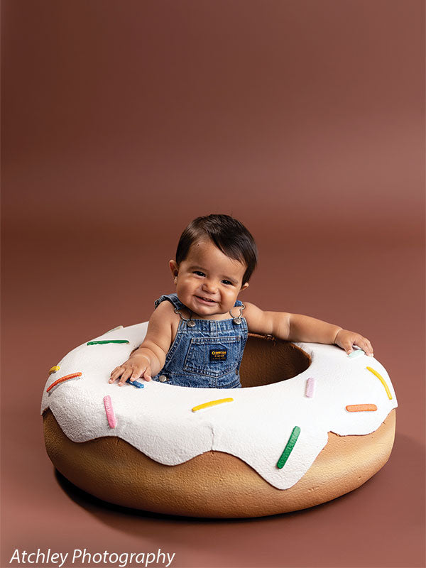 Baby wearing blue denim overalls sitting inside a large donut prop with white icing and colorful sprinkles, set against a solid warm brown studio backdrop, smiling and resting both arms on the edge of the donut.