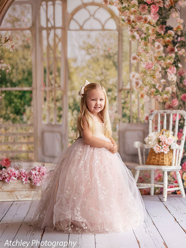 A young girl wearing a light pink tulle dress stands turned slightly to the side, smiling, with a floral conservatory backdrop featuring tall windows, blooming flowers, and soft greenery.