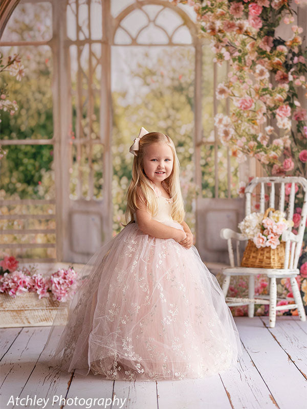 A young girl wearing a blush pink tulle dress stands turned slightly to the side, smiling, posed in front of a spring floral conservatory backdrop with tall windows, flowers, and greenery.
