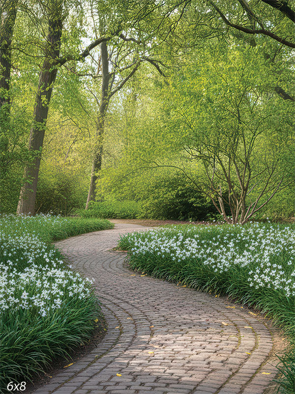 A winding cobblestone path lined with clusters of bright white spring flowers and lush green foliage under a soft tree canopy.
