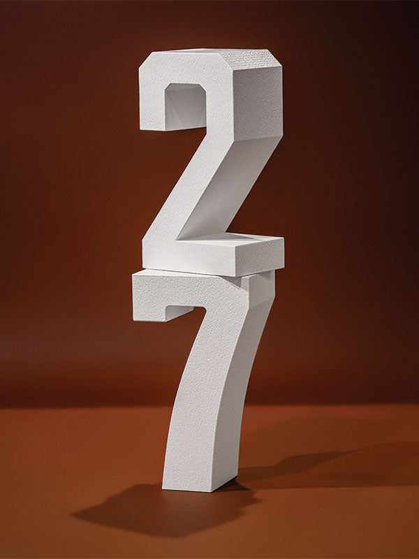 White hard-coated foam stand-alone number props stacked with a 2 balanced on top of a 7 on a warm orange background.
