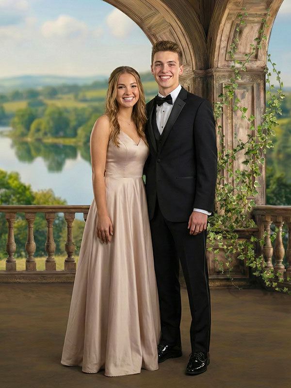A young couple stands together beneath a stone arch balcony backdrop overlooking a calm lake and rolling green countryside, with the woman wearing a champagne-colored floor-length dress and the man dressed in a black tuxedo, both smiling and facing forward.