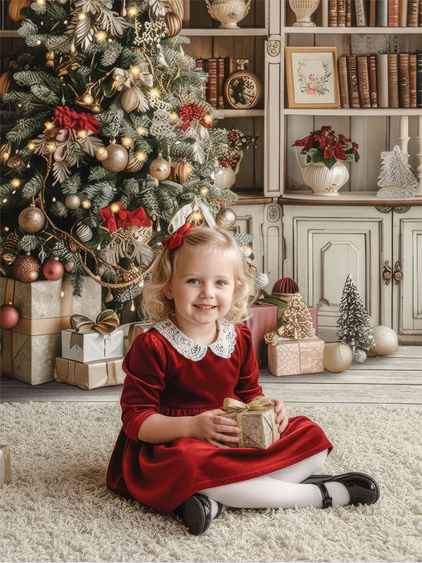 A young girl in a red dress sits on the floor holding a small gift in front of a decorated Christmas tree, posed against a cozy holiday library backdrop with bookshelves, wrapped presents, and festive accents.

