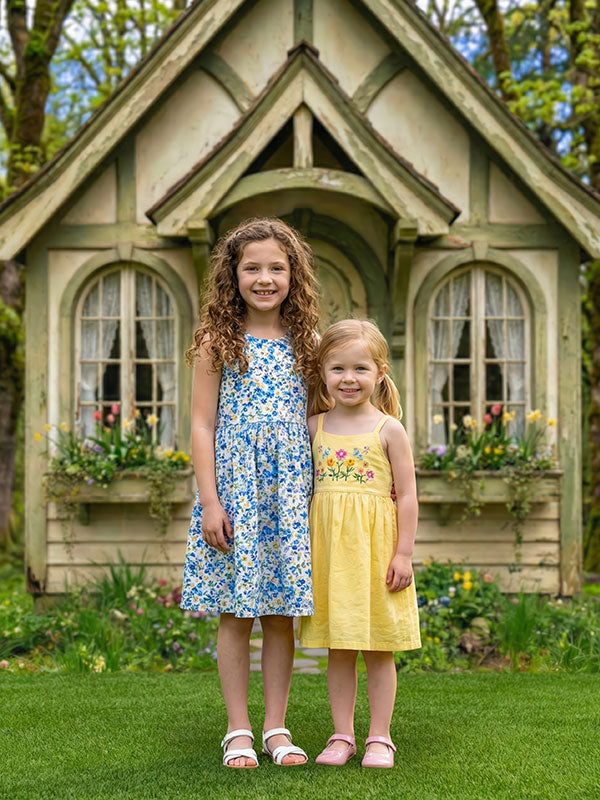 Two young girls wearing floral and yellow dresses stand side by side on grass in front of a charming storybook cottage garden backdrop with flower boxes, windows, and spring greenery.