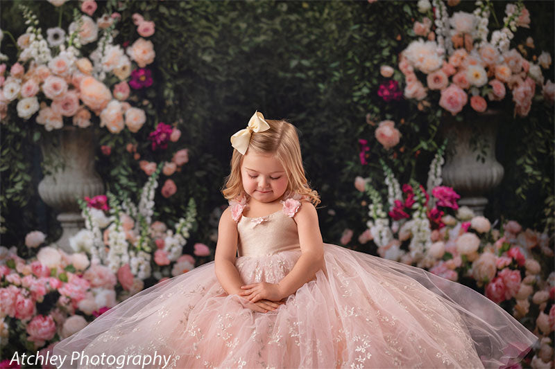 A young girl in a blush pink tulle dress sits gracefully with hands folded, posed against a storybook-style floral garden backdrop filled with roses and greenery.
