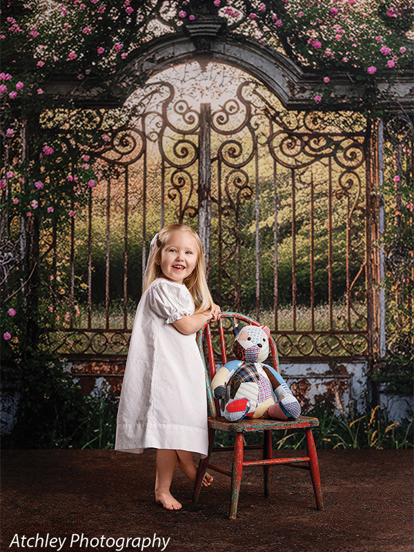 A young girl wearing a white dress stands barefoot beside a small chair holding a teddy bear, posed in front of a storybook-style garden gate arch backdrop with greenery and flowers.
