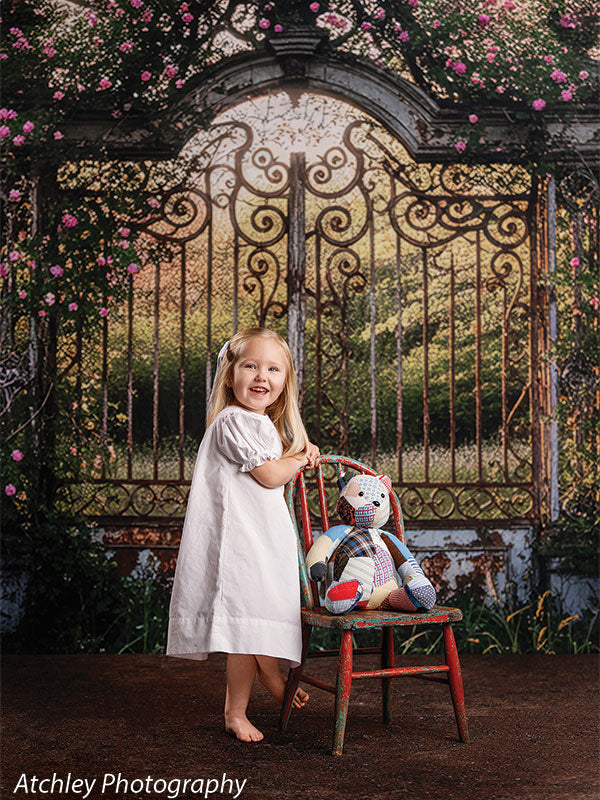 A smiling young girl wearing a white dress stands next to a small chair with a teddy bear, posed in front of a storybook-style iron garden gate backdrop with flowering vines.
