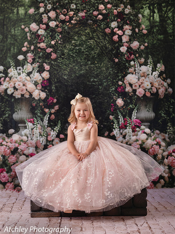 A young girl wearing a blush pink tulle dress sits centered with hands folded, posed in front of a storybook-style rose garden archway backdrop filled with flowers and greenery.

