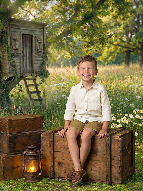 Young boy wearing a light shirt and brown shorts sits smiling on stacked wooden crates in front of a storybook outdoor field backdrop featuring a small treehouse, tall grass, daisies, and warm natural light.