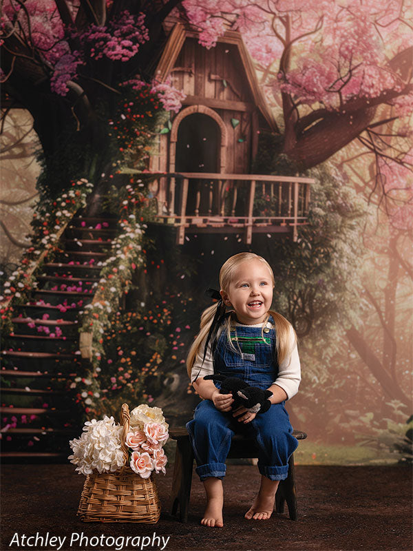 A young girl in denim overalls and a white shirt sits barefoot on a small chair holding a stuffed animal, smiling while looking to the side, posed against a storybook-style treehouse forest backdrop with flowers.
