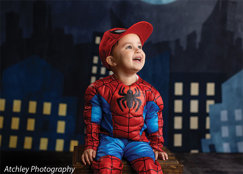 A toddler in a red and blue superhero costume and red cap sits on a wooden box looking upward, set against a stylized night cityscape backdrop with illuminated windows and a moon.
