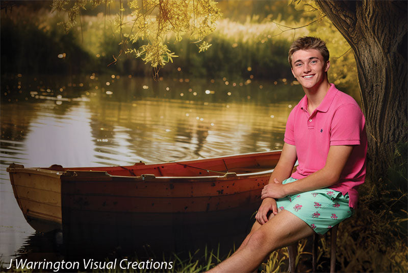 Teen boy smiling while seated beside a red wooden rowboat on a calm lake backdrop with golden sunset lighting and tree branches, ideal for summer-themed senior portraits.