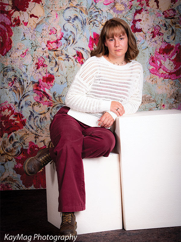 Teen girl with short bangs and a cozy outfit posing on white blocks in front of a rich, vintage-style floral tapestry backdrop, great for boho or editorial portrait styles.