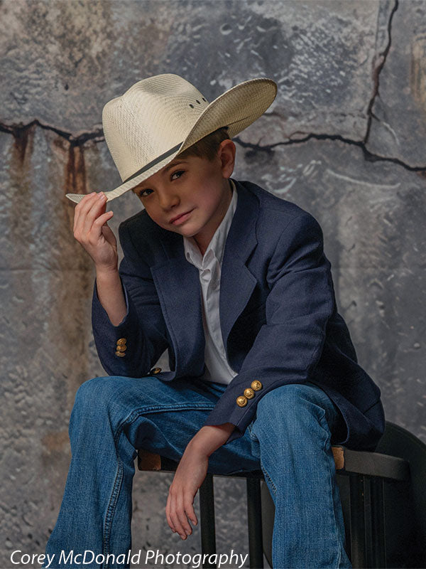 Young boy in a navy blazer, white shirt, and blue jeans sits on a bench while tipping a tan cowboy hat with one hand in front of a textured gray concrete wall backdrop with cracked urban detailing.