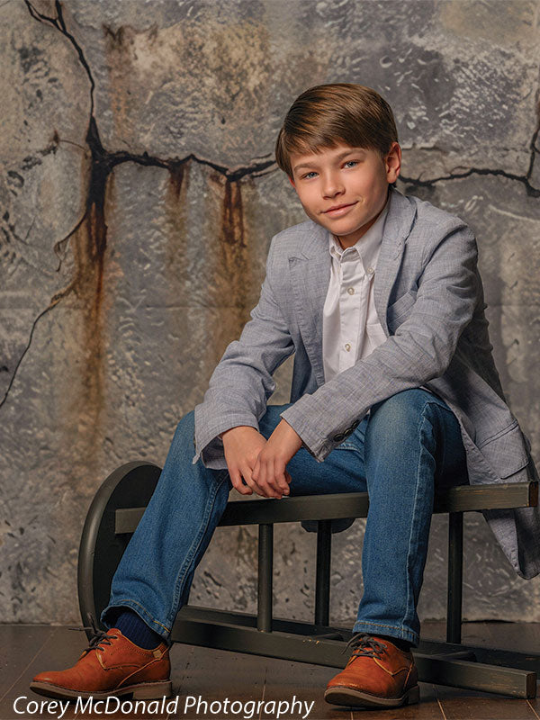 Young boy in a light gray blazer, white shirt, and jeans sits casually on a bench with hands resting together in front of a textured gray concrete wall backdrop with cracked urban detailing.