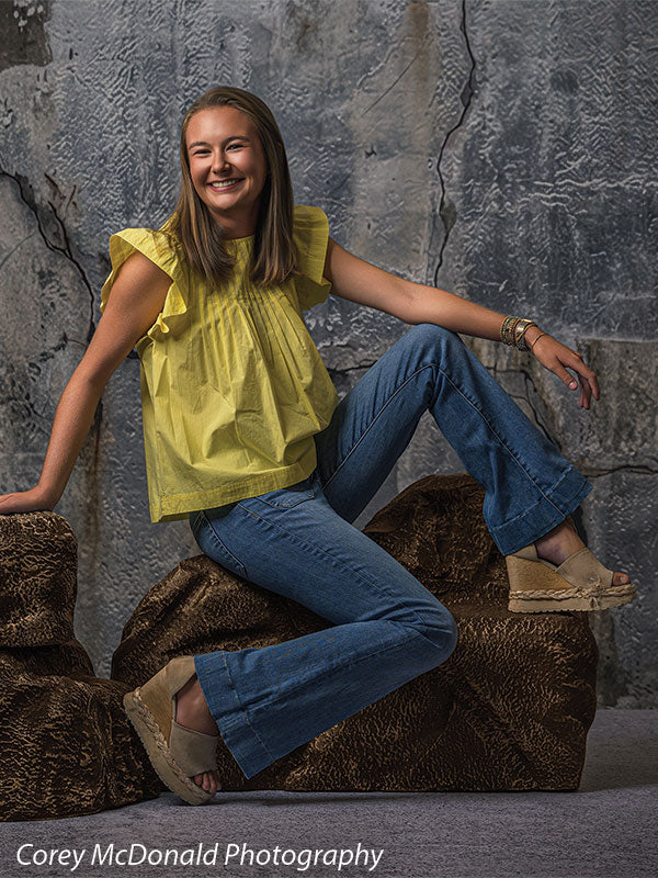 Textured Silver Industrial Wall photography backdrop shown with a senior girl in a yellow ruffle-sleeve top and blue flared jeans leaning back on the brown rocks with a wide smile on faux rock props CP-8807
