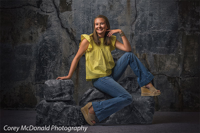 Textured Silver Industrial Wall photography backdrop shown with a senior girl in a yellow ruffle-sleeve top and blue flared jeans leaning on the grey rocks and smiling on faux rock props CP-8807