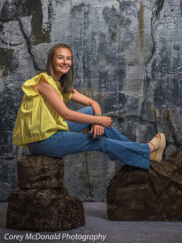 Textured Silver Industrial Wall photography backdrop shown with a senior girl in a yellow ruffle-sleeve top and blue flared jeans reclining low against the brown rocks on faux rock props CP-8807